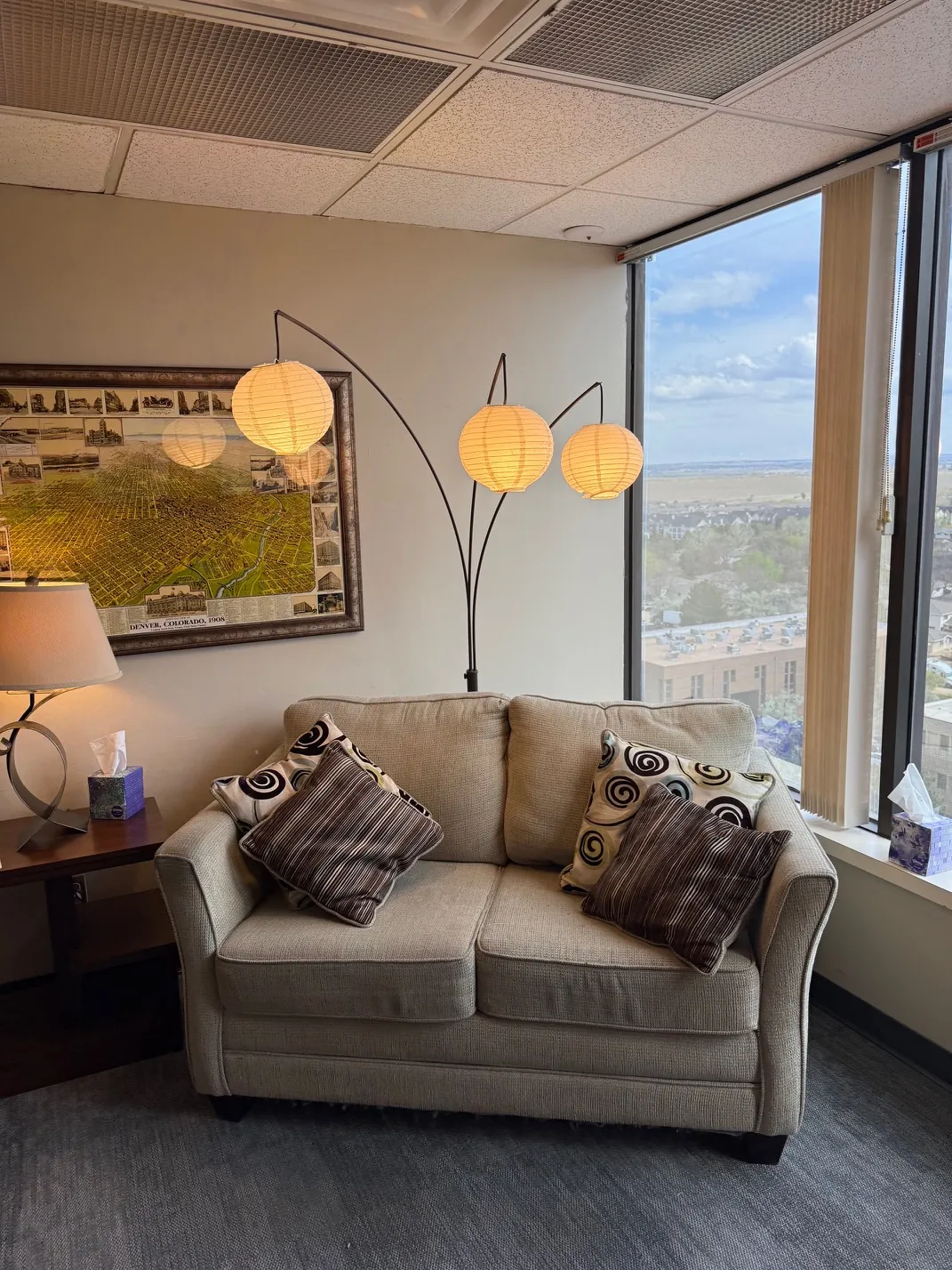 A quiet corner of the Colorado Center for Couples and Families office: a beige loveseat with patterned pillows beside a south-facing window with a Denver view, under a 1908 map of Denver and a tall arc lamp with paper lanterns.